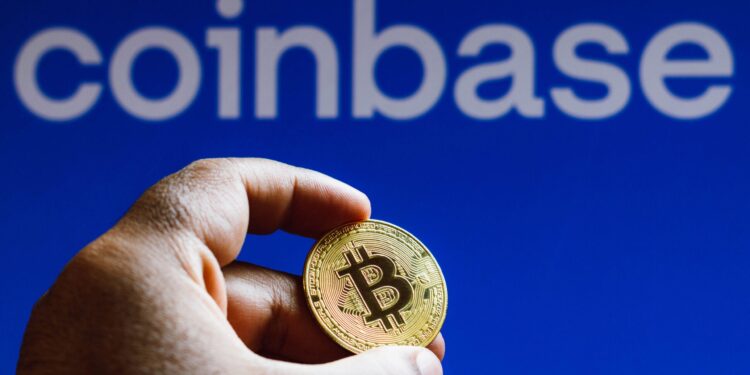 Una mano che tiene una moneta d'oro con il simbolo del Bitcoin davanti a un logo di Coinbase blu.