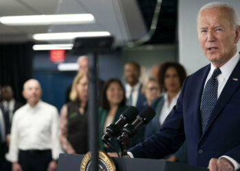 Joe Biden parla davanti ai microfoni durante un evento pubblico, con un gruppo di persone sullo sfondo