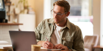 Image of focused young man in glasses, sitting in cafe, making notes, studying, attending online course, learning on remote from quite coffee shop.